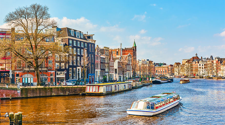 Canal tour boats and houseboats float past traditional gabled houses on a sunny day in Amsterdam.