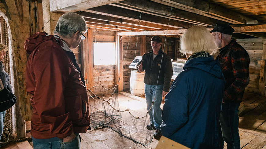 A tour guide gestures towards a fishing net while speaking to a group of travelers inside a rustic wooden building in Newfoundland and Labrador.