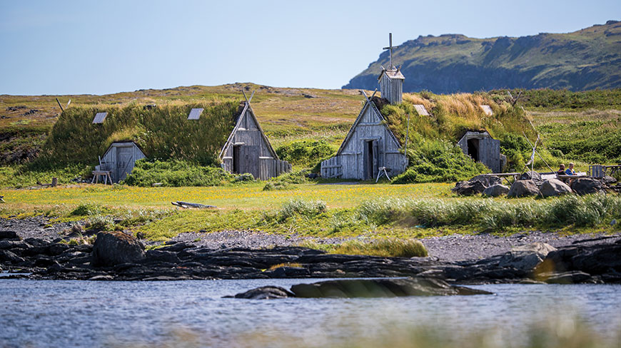 Several turf-roofed wooden huts in the Norstead Viking Village sit on a grassy shore next to the water in Newfoundland and Labrador.