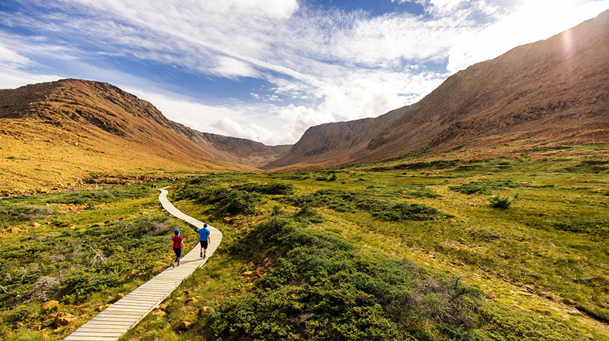 Two hikers walk along a wooden boardwalk through the golden, mountainous Tablelands of Gros Morne National Park in Newfoundland and Labrador.