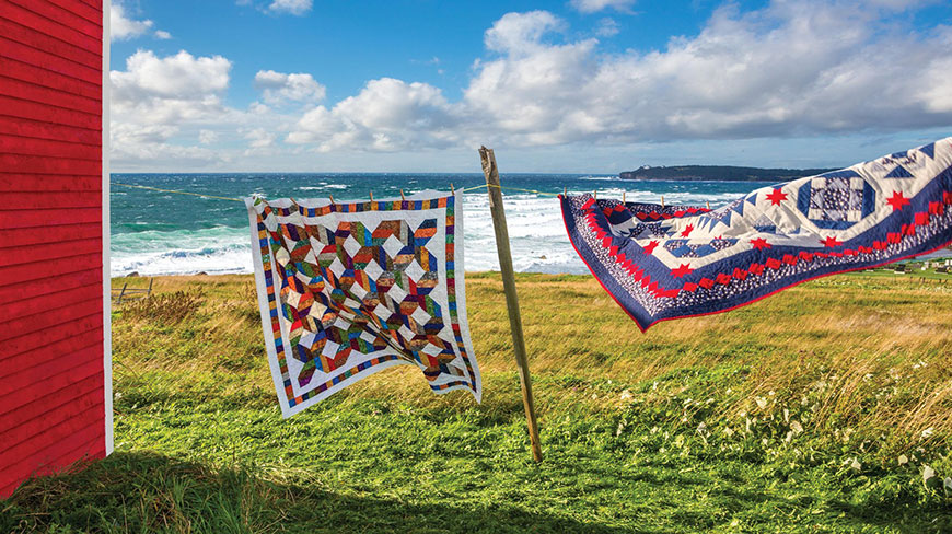 Two colorful quilts hang on a clothesline in a coastal field in Newfoundland and Labrador, with the ocean in the background.