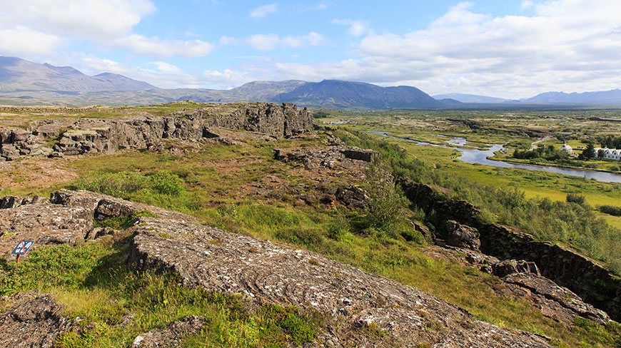 A view across the rocky, grass-covered rift valley of Þingvellir in Iceland, with a river and mountains visible in the distance.
