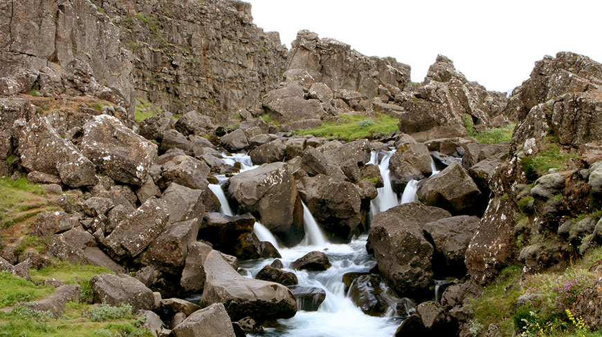 A small waterfall cascades over large, mossy boulders in a rocky gorge in Thingvellir, Iceland.