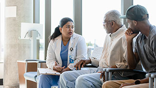 A female doctor discusses something on a clipboard with an older male patient and a younger man in a waiting room.