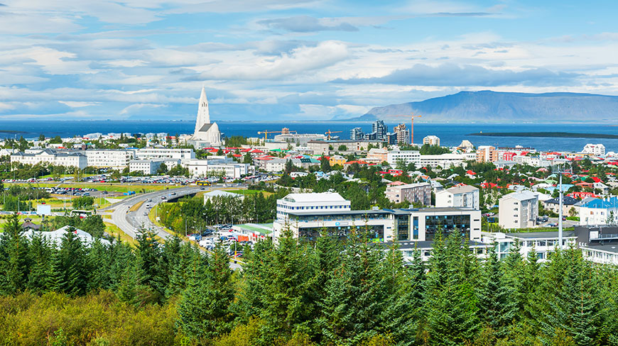 A high-angle view of Reykjavik, Iceland, shows the city stretching to the ocean, with lush green trees in the foreground.