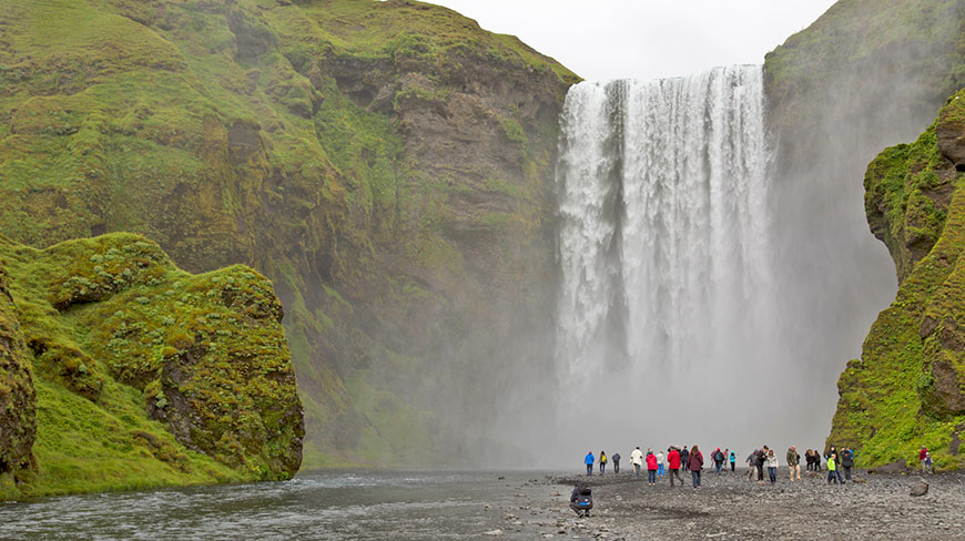 A crowd of people stands on the rocky bank at the base of the massive Skógafoss waterfall in Iceland, surrounded by green cliffs.