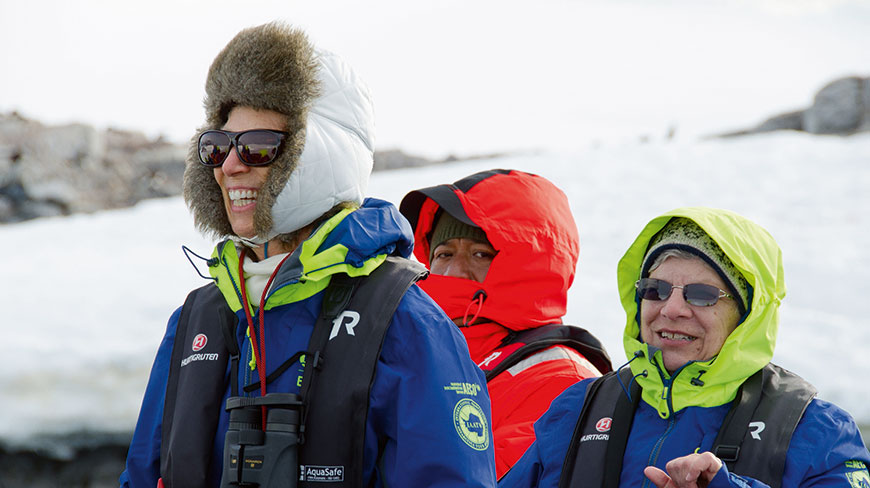 Three people in colorful parkas and life jackets ride in a zodiac boat through a snowy, arctic landscape.