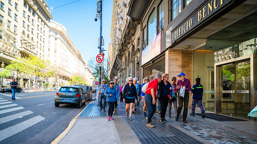 A group of travelers on a walking tour explores a city street in Buenos Aires lined with classic architecture on a sunny day.
