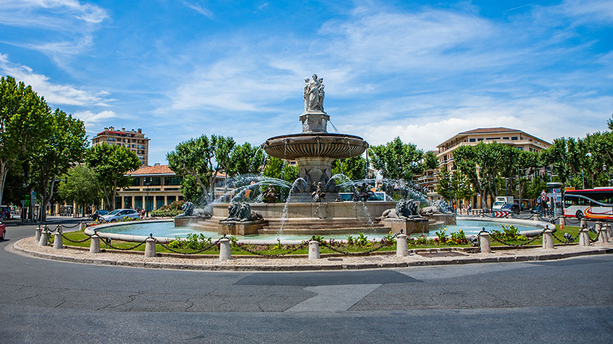 The Rotunda Fountain in Provence, France, sits in a roundabout surrounded by trees and buildings on a sunny day.