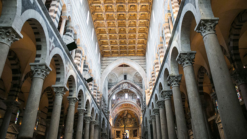 A low-angle view of the ornate golden ceiling, striped marble arches, and tall columns inside a grand cathedral in Pisa, Italy.