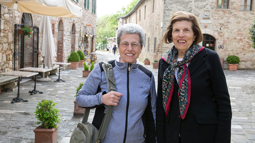 Two women smile while standing on a cobblestone street in a medieval village in Tuscany, Italy.
