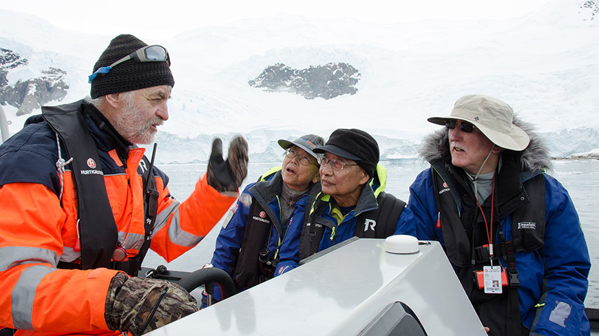 A guide in a bright orange jacket speaks to three passengers on a small boat in a glacial environment.