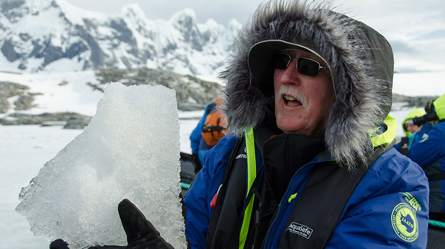 A man in a blue parka with a fur-trimmed hood holds up a large piece of ice in a snowy, mountainous setting.