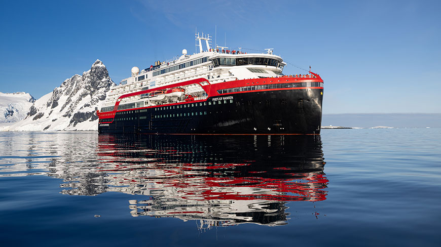 The MS Fridtjof Nansen expedition ship floats in calm water in Greenland, with snow-covered mountains rising from the sea behind it.