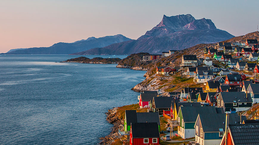 Colorful houses line the rocky coast of Greenland, with dark, jagged mountains rising in the distance across the water.