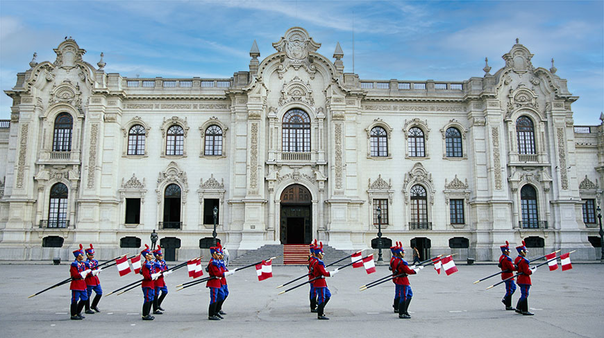 Soldiers in red and blue uniforms march with Peruvian flags in front of the ornate Government Palace in Lima, Peru.