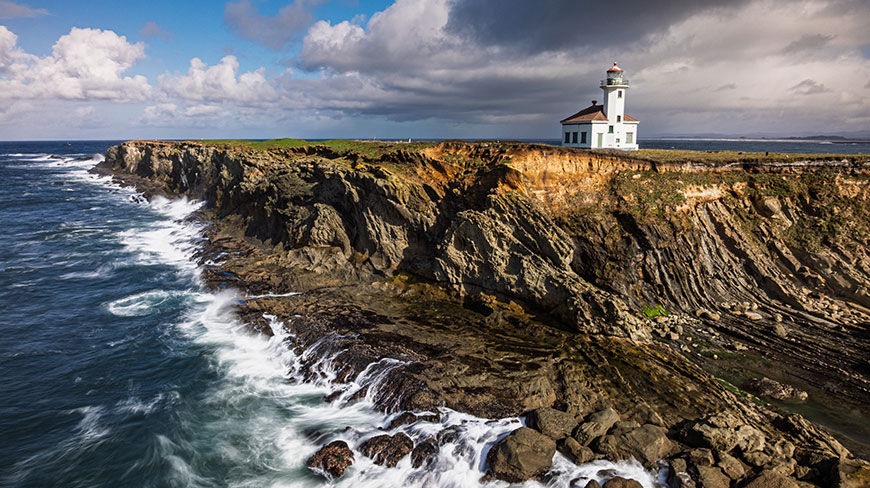 An aerial view of a white lighthouse on a rocky cliff overlooking the ocean with waves crashing on the shore under a partly cloudy sky.