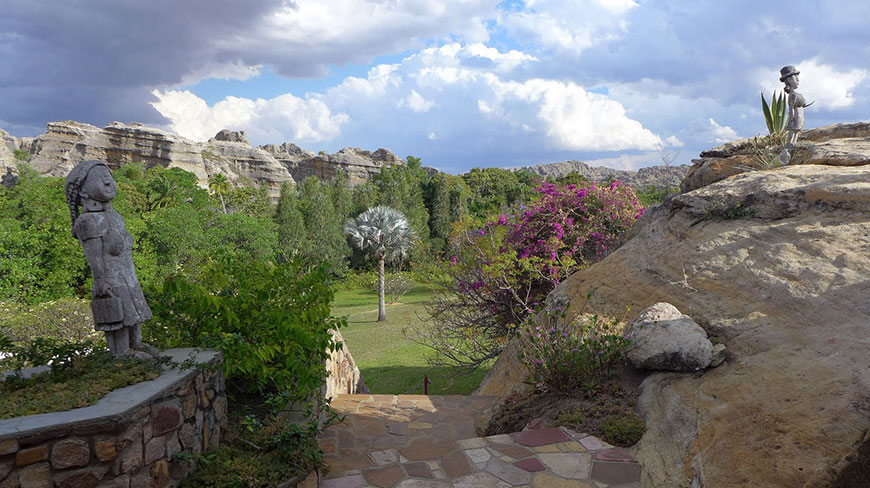 Stone steps lead down into a lush garden with unique statues, tropical plants, and dramatic rock formations in Madagascar.