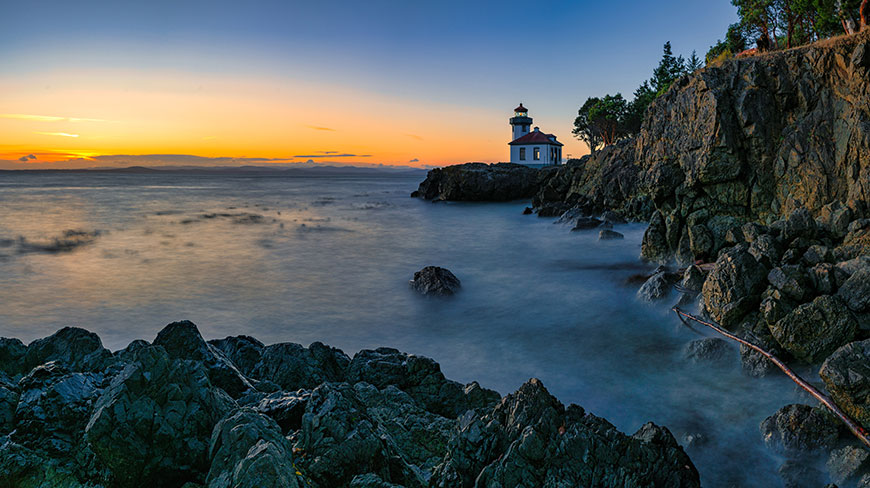 A white lighthouse sits on a rocky coast as the sun sets over the water, creating a golden glow on the horizon.