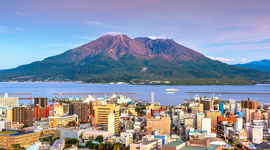 The cityscape of Kagoshima in Japan overlooks the bay with the active Sakurajima volcano in the background at sunset.