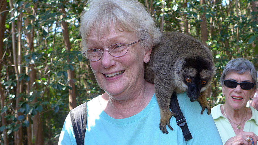 In Madagascar, a smiling woman has a brown lemur resting on her shoulder as another woman watches from behind.