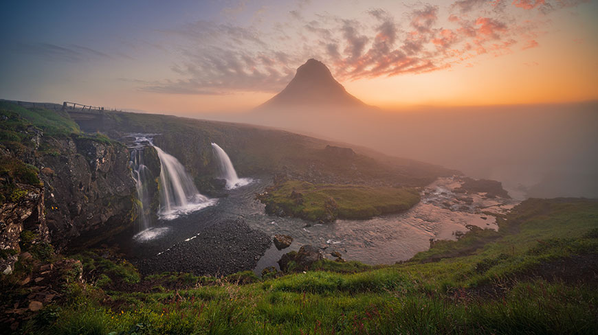 A dramatic, misty mountain peak rises above waterfalls cascading down a rocky cliff into a river during a colorful sunset.