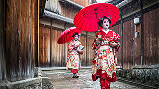 Two geishas in traditional red kimonos hold matching red umbrellas while walking down a narrow, traditional street in Japan.